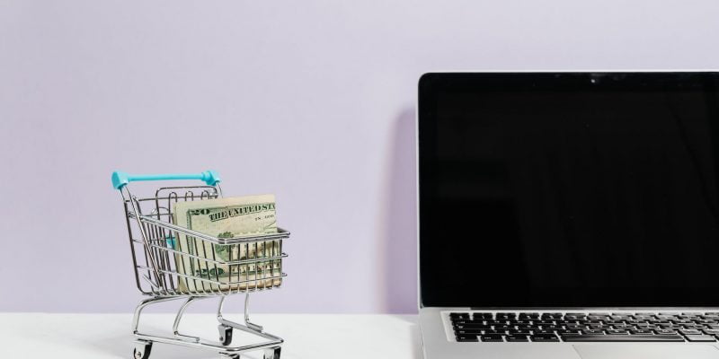macbook pro on white table beside a miniature shopping cart with money